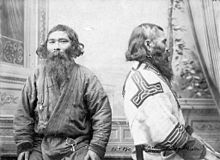 A black and white photograph of two Ainu men sitting next to each other, wearing traditional attire.